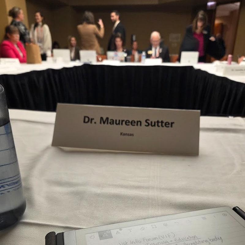 A conference table with a name card reading "Dr. Maureen Sutter, Kansas" in focus. People are gathered and conversing in the background, with papers and a water bottle on the table in the foreground.