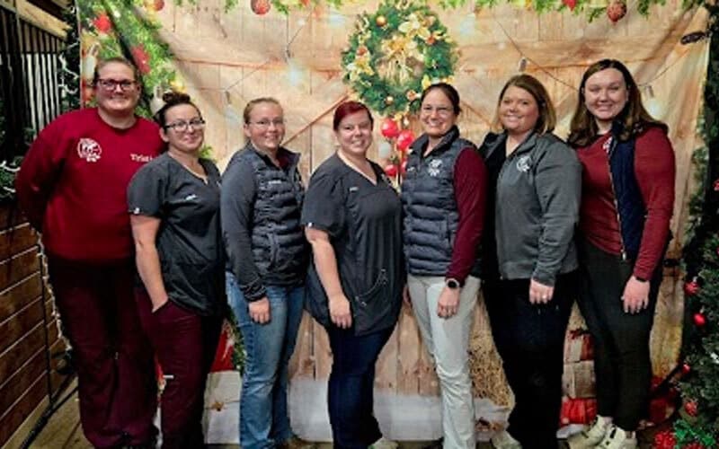 Seven women stand side by side, smiling in front of a festive Christmas backdrop decorated with a wreath, ornaments, and holiday lights. Some wear matching uniforms, while others wear casual attire.