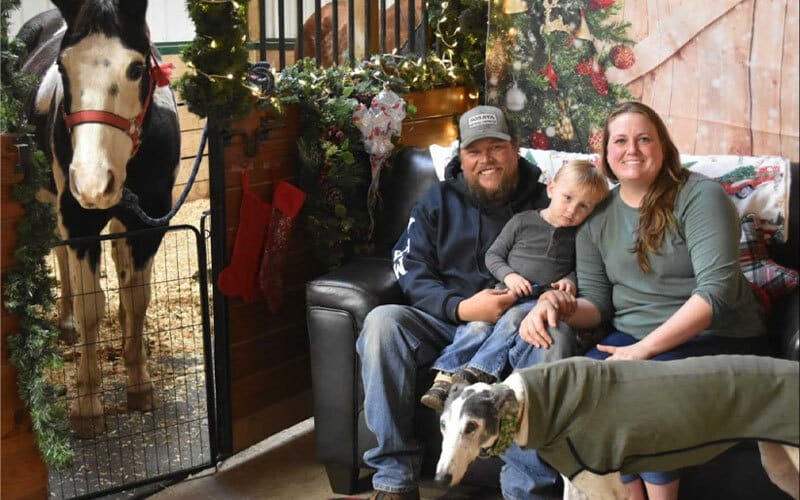 A smiling family with a young child sits on a couch indoors, surrounded by Christmas decorations. A horse stands in a nearby stall, and a dog wearing a coat is in the foreground.