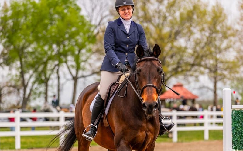 A woman wearing a helmet and riding jacket is riding a brown horse in an outdoor equestrian arena with a white fence and trees in the background.