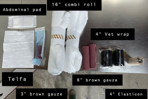 A medical supplies kit is laid out on a table. Items include an abdominal pad, 16" combi rolls, 4" vet wraps, Telfa pads, 3" and 6" brown gauze rolls, and a 4" Elasticon, each labeled with black tags.