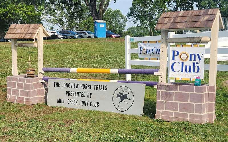A horse jump with stone-like posts and wooden roofs stands on grass. A sign reads “The Longview Horse Trials Presented By Mill Creek Pony Club.” Pony Club banners are visible on a white fence in the background.