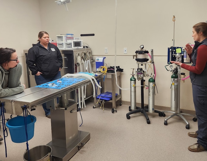 Three women are in a veterinary clinic exam room with medical equipment, oxygen tanks, and a metal examination table. One woman is explaining equipment while the others listen and observe.