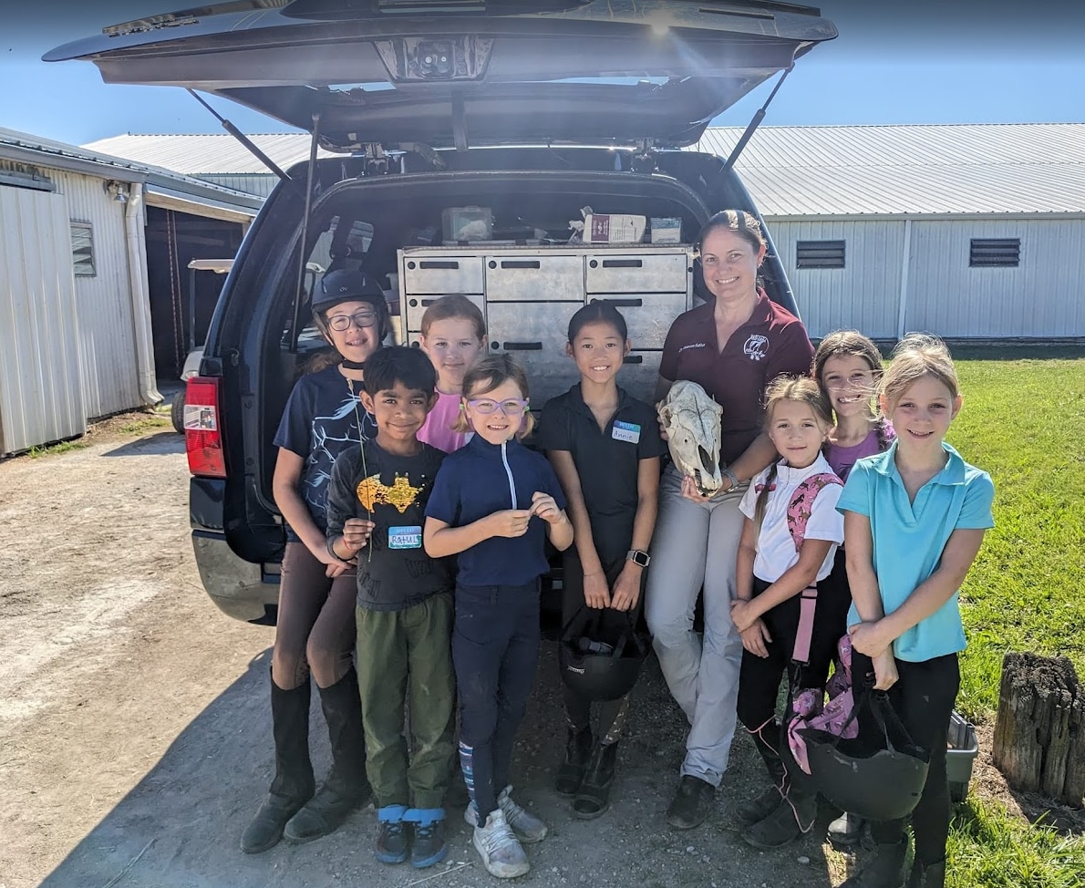A group of smiling children and one adult stand in front of an open vehicle filled with equipment, outside near barns. Some children hold riding helmets, and the adult holds a horse skull. All are dressed casually.