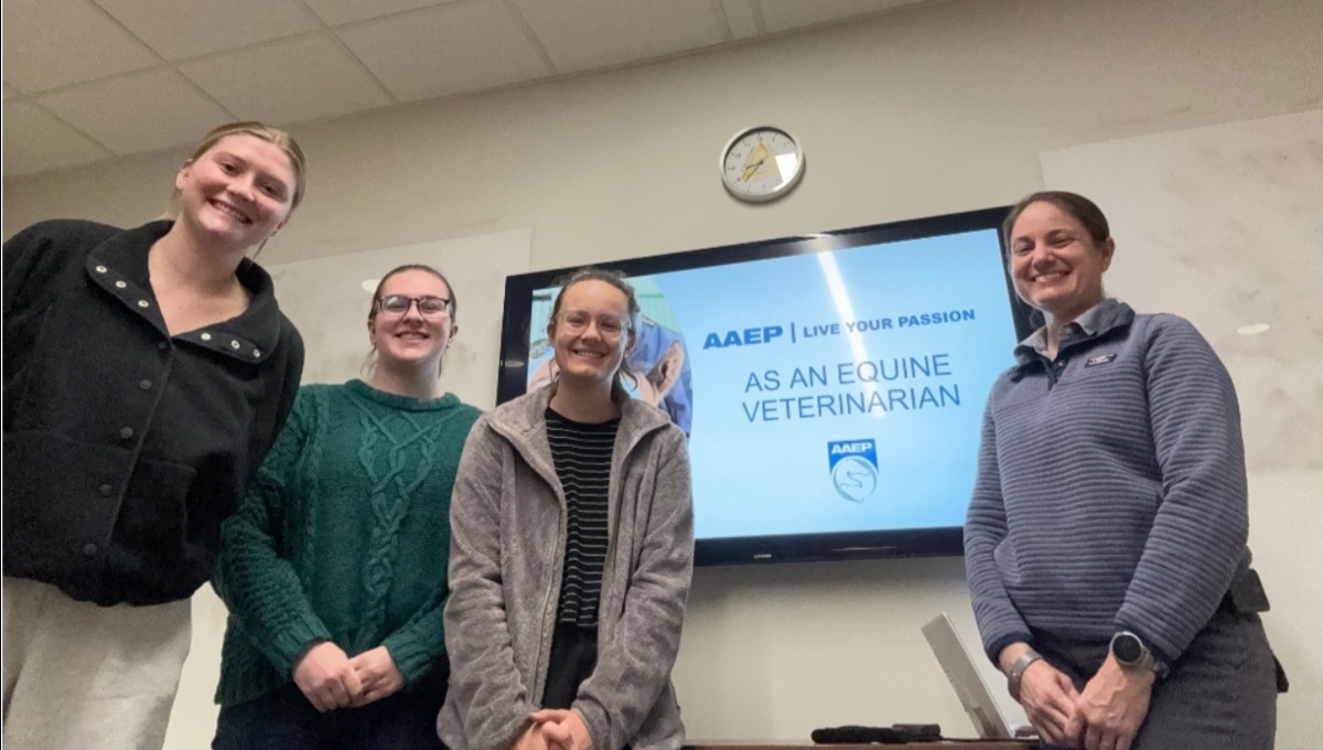 Four smiling people stand in front of a TV displaying a slide that reads, "AAEP LIVE YOUR PASSION AS AN EQUINE VETERINARIAN," with the AAEP logo. A clock is mounted on the wall above the screen.
