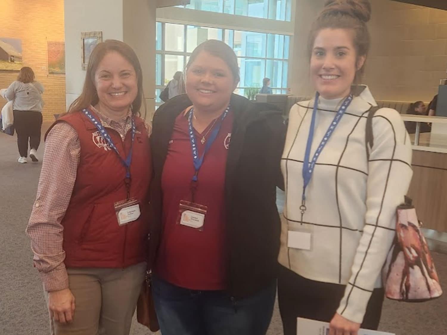 Three women standing indoors, smiling at the camera. Two are wearing maroon shirts with name badges and blue lanyards, while the third wears a white sweater with a grid pattern and carries a bag. The background shows a modern, open space.