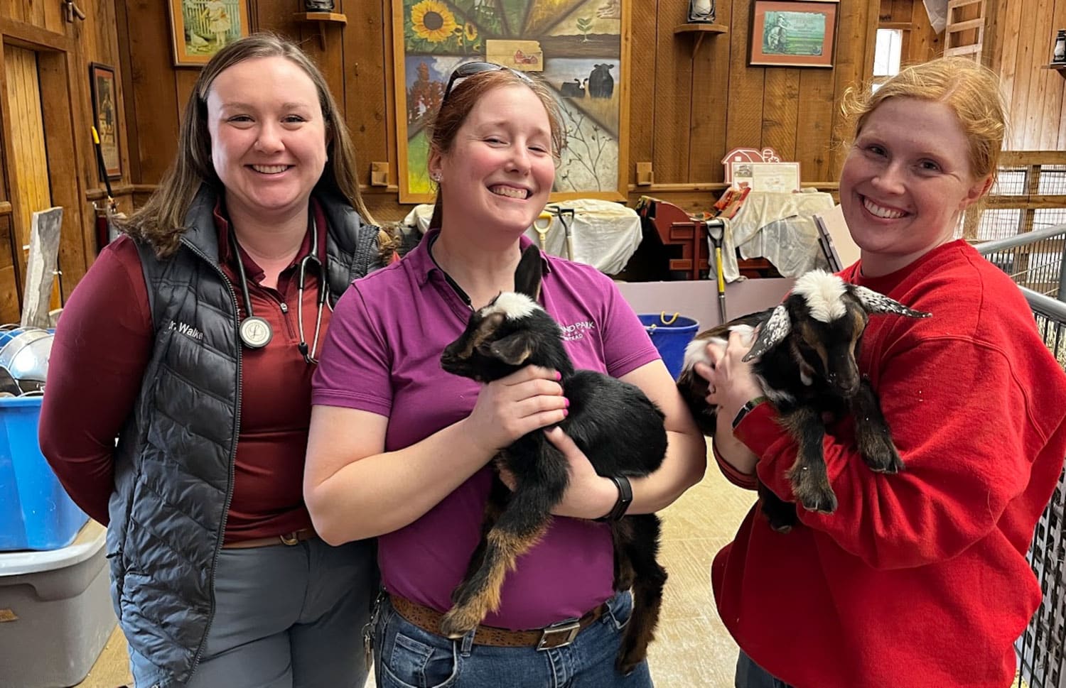 Three women smiling indoors, each holding a young black-and-brown goat. The background features wooden walls, paintings, and various farm or veterinary equipment.