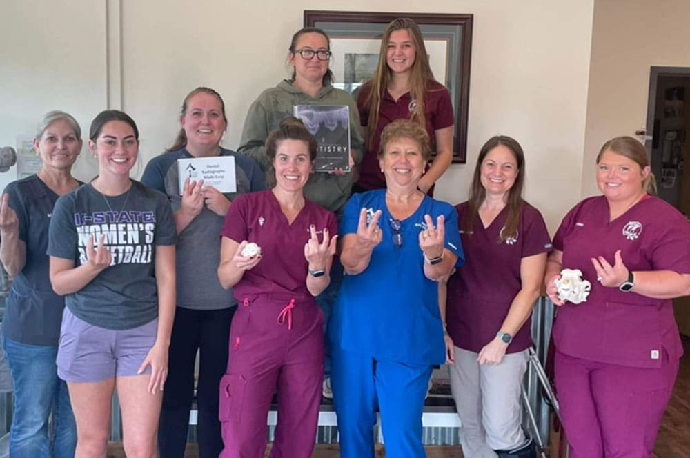 A group of nine women, some in medical scrubs and others in casual clothes, stand and smile indoors. A few are holding papers or items, and several are making hand signs with their fingers.